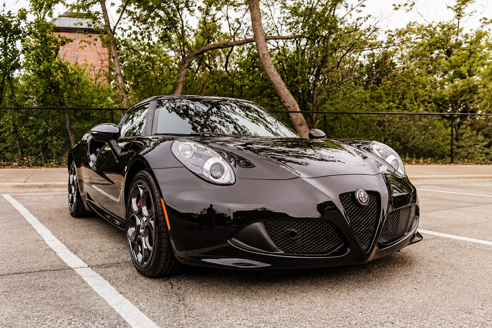 A black Alfa Romeo 4C parked on asphalt with trees and bushes behind it.